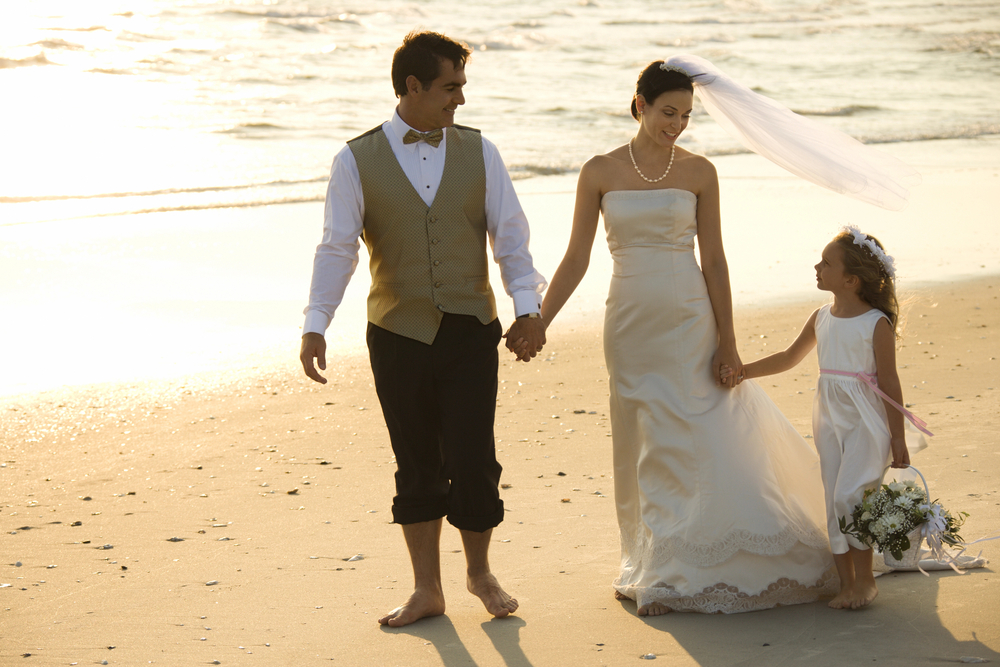 Little girl in dress walking with bride and groom on beach