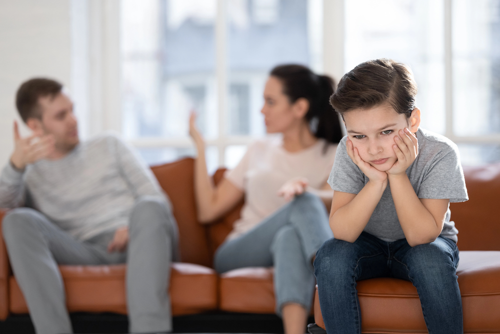 Upset boy sitting on couch with parents fighting in background