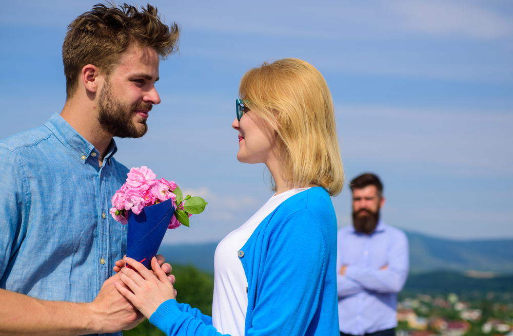 Man giving woman bouquet of flowers while woman's ex stands in background
