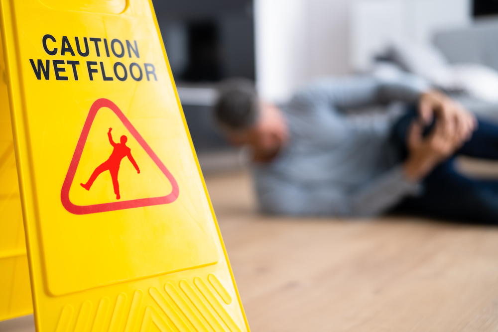 Close up of wet floor sign with man on floor in the background, clutching injured leg