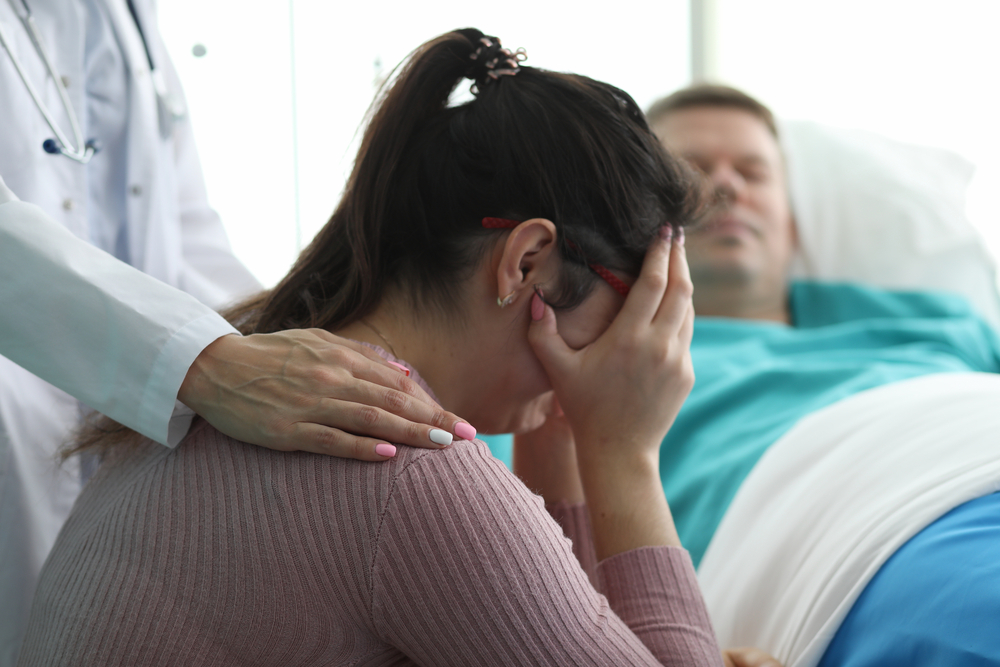 Woman crying next to husband's hospital bed with doctor's hand on her shoulder