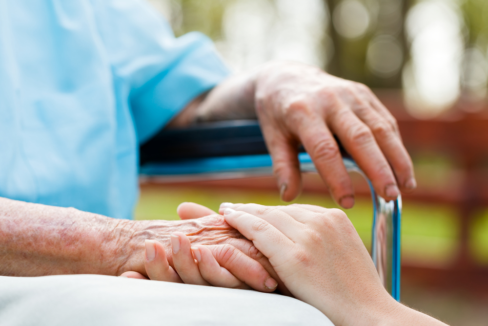 A younger hand holding an elderly hand extended from someone in a wheelchair outdoors