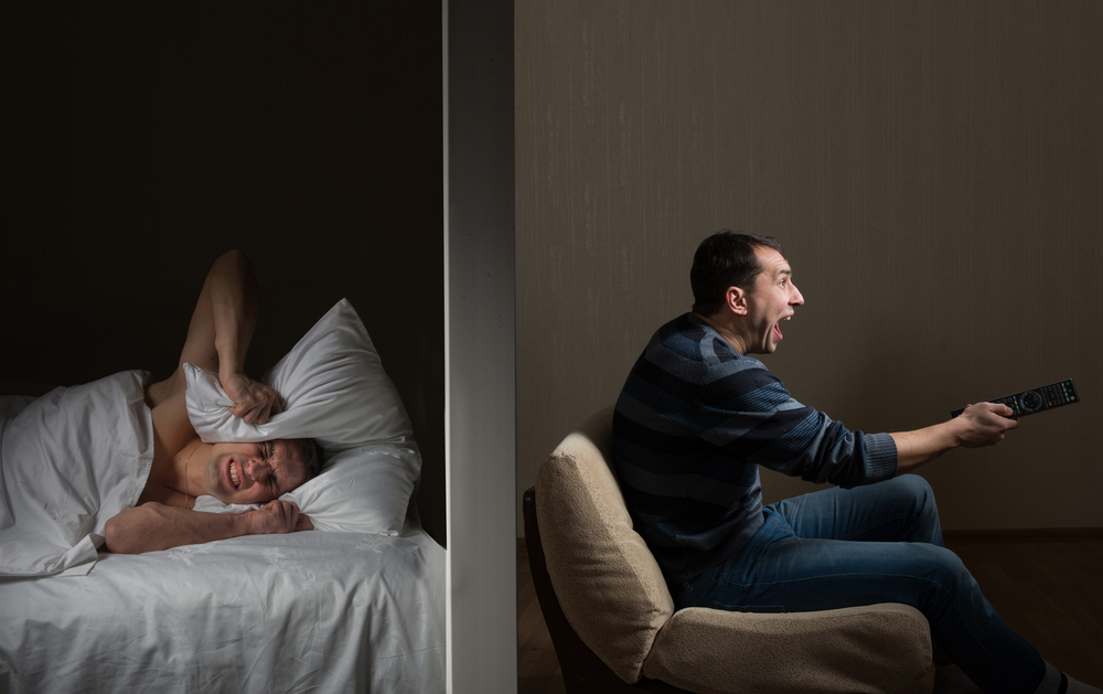 Man covering head with pillow while neighbor yells at TV